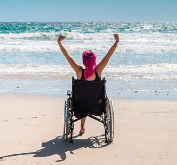 mujer en silla de ruedas de frente al mar mirando una ola y levantando ambas manos con sus puños cerrados